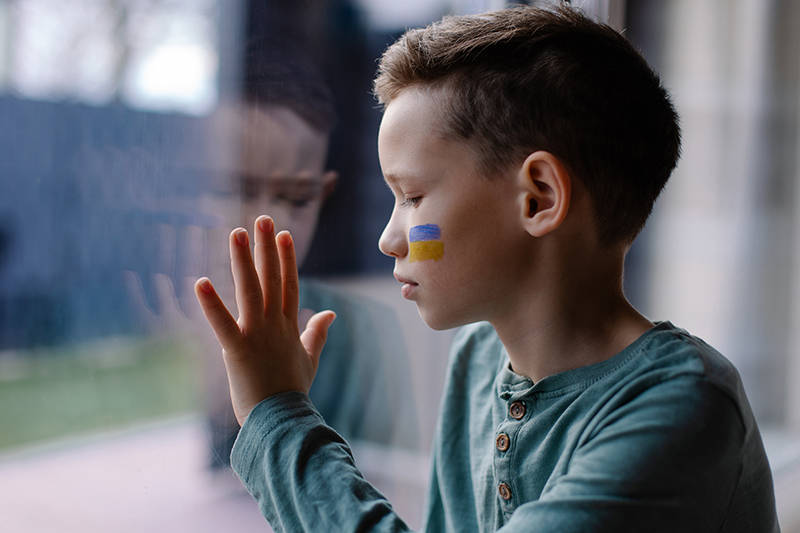 Image of a young boy with an Ukranian flag painted on his face looking out through a window
